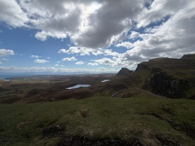 A landscape image shows an elevated view of a landscape in Portree, United Kingdom. Three lakes are visible in the distance. A winding road is visible on the right side of the image. Rolling hills and cliffs extend into the distance. The foreground is composed of a grassy, mossy terrain. The sky is partially cloudy with patches of blue sky peeking through. The sun is visible, though diffused by clouds.