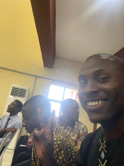 A selfie captures a group of four people in an indoor setting with yellow walls. The smiling individual at the top is wearing a dark shirt. Another person is wearing a shirt with a circular pattern. An air conditioning unit is on the wall in the background. A person in a white shirt and tie is sitting at a table. Someone is making a peace sign. The image is angled and slightly blurry.