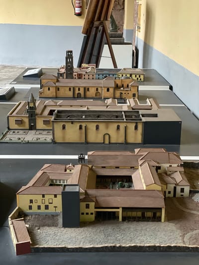 This media file presents an intricately detailed architectural model of a city, likely San Cristóbal De La Laguna, Spain, viewed from an elevated, slightly oblique perspective. The model features numerous buildings constructed from materials resembling light beige or yellow walls with reddish-brown tiled roofs, typical of historical European architecture. Many structures also incorporate dark flat roofs, suggesting different architectural styles or modern additions within the representation.
A prominent feature is a tall, dark tower with several levels and arched openings, standing amidst the densely packed miniature buildings. Streets are depicted in a dark grey color, with lighter grey elements possibly representing sidewalks or road markings. Other areas within the model show textured ground, some appearing cobbled or unpaved. The scale of the model suggests a significant level of detail, showcasing a complex urban layout with various building types and configurations, including courtyards and open spaces.
In the background, outside the model's immediate area, an indoor setting is visible. This includes a yellow painted wall, a grey painted wall, and some wooden structures or beams. A red fire extinguisher with unreadable white text is affixed to the yellow wall. No specific activity or event is taking place within the static model; it serves as a visual representation of the city's architecture and layout. The lighting appears to be artificial, indicative of an exhibition or display setting.