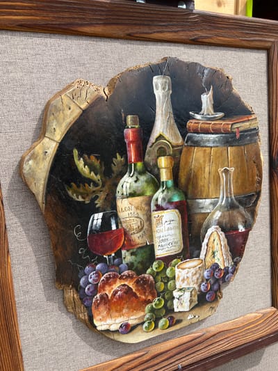 This is a painting of a still life on a piece of wood, framed by two wooden planks, and set against a textured grey fabric background. The painting depicts a rustic arrangement of food and drink. In the foreground, there are grapes, bread, and several pieces of cheese, including a wedge of blue cheese. Behind the cheese, there are two bottles of wine, one green and one red. To the right of the bottles is a wooden barrel. Further back, there are stacked books, a lit candle, and another bottle. The overall scene evokes a sense of abundance and tradition. The painting is signed "MB" in the lower right corner. There is no information in the image to suggest a specific location in the city of Yakunino, Russia.