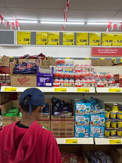 A person wearing a red hooded sweatshirt and a blue baseball cap is standing in a supermarket aisle. The aisle is stocked with various confectionery products such as Kinder Joy eggs, Milikta Bites, Yupi sweets, Delfi chocolates, and Cadbury Dairy Milk chocolate. Price tags with the local currency (Rp) are visible next to the products, indicating the cost of each item. The supermarket aisle is well-lit, and red and white bunting is hung along the top.
