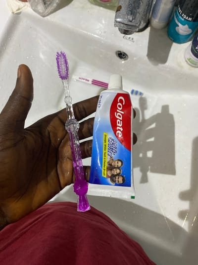 The image captures a close-up, eye-level view of a person's dark-skinned hand holding a tube of Colgate toothpaste and a purple toothbrush over a white bathroom sink. The hand appears wet, suggesting recent use of water or preparation for hygiene.

The main subjects are the hand, the Colgate toothpaste, and the purple toothbrush. The toothpaste tube is white and blue, featuring the "Colgate" logo prominently in red. Below the logo, it reads "ANTI-CAVITY" with the tagline "Stronger, Whiter Teeth & Fresher Breath." A small image of a family (a woman, a child, and a man) is visible on the tube. The volume "75 mle" is also printed on it. The purple toothbrush, held horizontally beneath the toothpaste tube, has clear plastic bristles and a patterned handle.

In the background, within the sink basin, several other toiletries are visible. To the left, a silver faucet shows signs of wear, and next to it are bottles, including one partially visible with "AVE FOAM" printed on it, likely shaving foam. Another toothbrush, pink and white with "REACH" visible on its handle, lies in the sink to the left of the hand. The sink itself is white and appears wet, with some water droplets and possibly soap residue. A dark drain hole is visible in the center of the sink. Strong overhead lighting casts distinct shadows of the bottles and other objects onto the sink's surface, creating a somewhat stark contrast. On the far right of the frame, a portion of a red garment is visible, likely clothing worn by the person holding the items.

The scene depicts a common morning or evening routine, likely in a residential bathroom in Lakka, Sierra Leone, as indicated by the context. The items are arranged as if about to be used for dental hygiene.