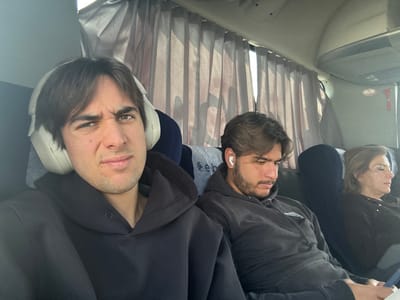 Three people sit on a bus with closed curtains. One person faces the camera, wearing headphones. Another has an earbud in and looks to the side. The last person is sleeping and is to the left. All are in darker clothing.