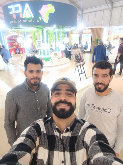 This indoor photograph captures a selfie of three men in the foreground, smiling at the camera, within what appears to be a brightly lit exhibition or trade show booth. The man in the center foreground wears a black and white plaid shirt and a beige baseball cap with a dark "LA" logo. To his left, another man wears a grey button-up shirt, and to his right, a man in a light grey sweatshirt featuring the reversed text "Calvin Klein collection". All three have beards.
The background reveals a bustling event space. A large, circular, dark blue overhead sign prominently displays the light-up letters "AFC" and the phrase "A vision to share" (appearing reversed due to the selfie perspective), alongside a colorful, stylized map of Africa. The booth features modern design elements, including hanging artificial plants, neon strip lighting, and display shelves stocked with various products, possibly food items or packaged goods. QR codes are visible on some stands. People are scattered throughout the background, sitting at tables, conversing, and exploring the exhibits. A notable detail is a framed portrait on an easel, depicting a formally dressed man (likely a monarch, given the context) with Arabic script below it, adding a cultural cue to the scene. The atmosphere appears lively and engaging, consistent with a public or professional event.