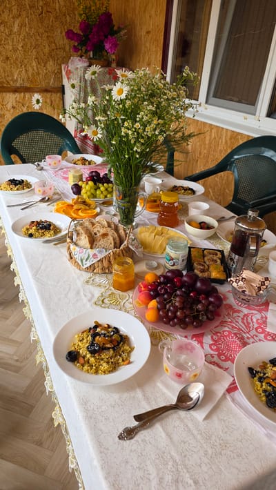 A table covered with a white tablecloth is set for a meal in what appears to be an outdoor or semi-outdoor setting, possibly a veranda or gazebo. The background is a wooden wall. On the table, there are several plates of food, including what looks like oatmeal or porridge topped with dried fruits and nuts. There are also bowls of fruit, including grapes and apricots, sliced oranges, a basket of bread, jars of jam, a container of coconut oil, and a French press coffee maker. A bouquet of daisies is prominently displayed in a vase in the center of the table. The table is also adorned with a decorative runner. Two green plastic chairs are visible on either side of the table. The lighting suggests it is daytime. There are no people visible in the image. The scene depicts a leisurely breakfast or brunch.