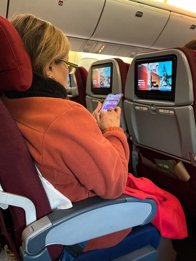 The image shows a passenger on an airplane, likely a Boeing 767-300 based on a partially visible tag. The individual, a woman with blonde hair, is wearing glasses and a dark-colored scarf or turtleneck under an orange fleece jacket. She is reclined in her seat, resting her head on a red headrest, and looking at a smartphone in her hands. The phone screen is illuminated with a display showing some text and icons. The in-flight entertainment system screen of the seat in front of her is displaying a welcome message, likely in Spanish given the word "Bienvenido". A red blanket is visible next to her, and a blue seat cushion is present. A partially visible armrest is situated to her right. The airplane's interior wall is visible in the background, with overhead compartments above. Given the presence of the Boeing 767-300 tag, it is possible the airplane is departing from or arriving at Arturo Merino Benítez International Airport in Pudahuel, Chile, but the image does not explicitly confirm this location.