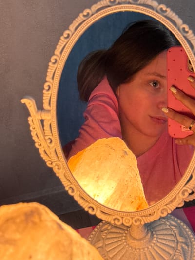 The media file is a close-up, horizontal, eye-level shot of a mirror reflecting a person. The reflection shows a person's face, with their head tilted and their left eye looking towards the camera. They are wearing a pink top and have dark hair. One hand, adorned with glittery nail polish and a ring, is visible at the top of the frame, holding a pink phone case with small red dots. The person's expression appears relaxed or contemplative.
The mirror itself is oval-shaped with an ornate, white frame featuring scrollwork and beaded details. The mirror is positioned in front of a wall that appears to be dark blue in the reflection, and a textured, light-colored object, possibly a lamp or a salt lamp, is visible to the right of the reflection. The overall lighting is warm, suggesting an indoor setting, possibly at dusk or night. No specific time of day, weather, or distinct location cues are evident beyond the indoor setting. No visible text is present in the media file.