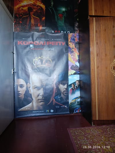This media file shows a vertical poster for the film "Kopoli Repu" displayed on a wall. The poster features a close-up of a man's face wearing a crown, with a woman's face visible in the background. Text on the poster indicates it is an information partner of "Nashe Radio" and that the film is showing in cinemas from August 3rd. Below the main poster are smaller images depicting scenes from other films, including a car and several people. The poster is mounted on a grey wall next to a wooden wardrobe. A patterned rug is visible on the floor. The timestamp "26.06.2026 12:10" is superimposed on the lower right corner of the image. The scene appears to be indoors, possibly in a room in Novovolynsk, Ukraine, given the provided location context. The lighting suggests daytime. The overall presentation suggests a promotional display for film releases.