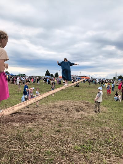 In Кояново, Russia, during an outdoor event, a woman in a blue dress is balancing on a long, thick log positioned across a grassy field. The log is angled, with its higher end supporting the woman. In the foreground, a young girl with blonde braided hair stands on the left, facing away from the camera and towards the event. Several children and adults are scattered throughout the background, with tents and structures visible in the distance, suggesting a festival or fair. The sky is overcast with dramatic clouds. The ground is covered in dry grass and some scattered hay.