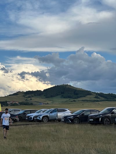 The image captures a scenic view of rolling green hills under a dramatic sky with a mix of blue patches and heavy, textured clouds, some illuminated by sunlight. In the foreground, a line of parked cars, primarily SUVs and sedans, are visible on a grassy field. A man wearing a white t-shirt with the number "045" on it, dark shorts, and a cap is walking across the field, facing away from the camera. Behind the cars, the landscape transitions to a densely forested hillside, leading up to a mountain peak that features a building or structure on its summit. Further in the distance, there are scattered buildings and structures nestled into the landscape on the lower slopes. The overall impression is one of a rural or semi-rural outdoor setting, possibly a gathering or event, given the presence of multiple vehicles and a person in athletic attire.