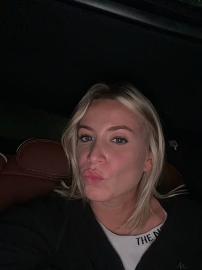 A blonde woman with light skin, blue eyes, and pursed lips is positioned at an angle, leaning against what appears to be a brown leather car seat. She's wearing a white tank top with the words "THE NOR" visible on the neckline and a black jacket or sweater. The background is dark, suggesting low lighting conditions, possibly inside a car at night. Her hair has blonde highlights and is styled in a loose, wavy manner.