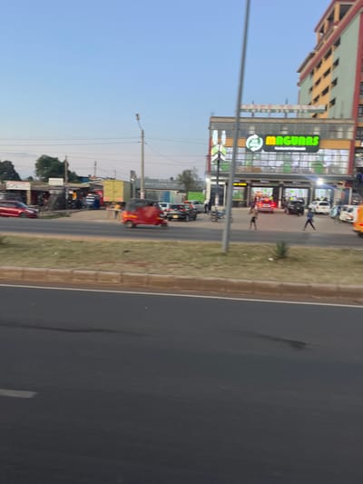 The image depicts a street scene in Biashara ward, Kenya, at dusk or early evening. In the foreground, a dark asphalt road is visible, bordered by a grassy median strip with a concrete curb. The midground features a busy street with vehicular and pedestrian traffic. A red tuk-tuk is prominently visible, traveling from left to right. Multiple other vehicles, including cars of various colors (red, dark, white) and a yellow truck, are present, some parked along the roadside, others in motion or waiting.

On the right side of the street, a two-story commercial building with a large green "MAGUNAS" sign and a smaller "24 HR M-PESA" sign is illuminated, indicating a retail establishment with significant window space. Above this building, a taller, multi-story structure with alternating yellow and green/red exterior panels is present. To the left of the Magunas building, a series of smaller, single-story commercial structures, some appearing as market stalls or shops, line the road. One sign appears to read "RESTORATION ZONE." Several pedestrians are visible on the pavements and near the Magunas entrance, with two individuals distinctly observed crossing or standing near the building's front. Utility poles and overhead wires are visible against a clear, twilight sky.