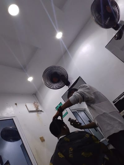 In Abeere, Nigeria, a barber is cutting the hair of a client in a dimly lit barbershop. Two ceiling fans are visible, along with overhead lights that illuminate the white walls and ceiling. The barber, wearing a light-colored sweatshirt, is using electric clippers on the client's head. The client, seated and facing away from the camera, wears a patterned cape. A mirror is partially visible in the background, and to the right, a sterilization unit is mounted on the wall. A door with a fan visible through its glass is on the left side of the frame.