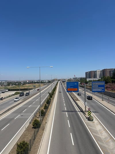 The image shows an aerial, wide-angle view of a multi-lane highway on a clear, sunny day. The highway stretches into the distance, with cars traveling in both directions. On the left side of the highway, there are tall, modern buildings, and on the right side, there is a grassy area with trees and a fence. Overhead streetlights line the highway.

Several blue highway signs are visible. One sign indicates destinations such as "Eceabat," "İstanbul," "Çan," and "Lapseki," along with a reference to "Karayolları 142.Şube Şefliği" (Highways 142nd Branch Directorate). Another sign points towards "18 Mart Üniversitesi Araştırma Hastanesi" (18 March University Research Hospital), "Kepez," "Şehir Merkezi" (City Center), and "Havaalanı" (Airport). A speed limit sign displays "50."

The sky is a clear, bright blue, suggesting it is daytime with fair weather. The overall scene depicts a typical urban or suburban highway environment, likely in Turkey given the text on the signs. There are no people prominently visible in the foreground, but cars indicate ongoing traffic. The composition of the image, with the highway disappearing into the horizon, gives a sense of depth and movement.