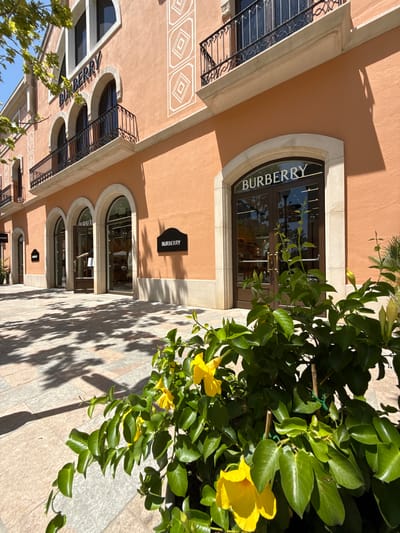 The image shows the exterior of a Burberry store in La Roca Village, La Roca del Vallès, Spain. The building has a peach-colored facade with arched windows and balconies with black wrought iron railings. The Burberry logo is visible above the main entrance, which features double glass doors. In the foreground, there are bright yellow flowers and green foliage in a planter, casting shadows on the patterned stone pavement. The sky is blue and visible through the leaves of a tree on the left side of the image. No people are clearly visible in the scene.