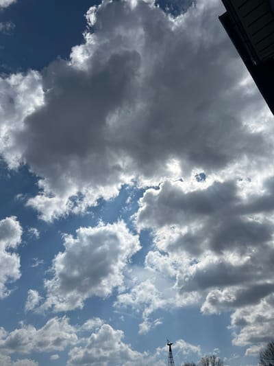 The image depicts a daytime sky over Byal Izvor, Bulgaria, dominated by a dynamic cloudscape. Large, dark grey cumulus and cumulonimbus clouds occupy the upper and central portions of the frame, interspersed with patches of clear blue sky. Bright sunlight penetrates through breaks in the cloud cover, creating visible light rays in several areas.

In the upper right corner, a dark, angular section of a building's eave or roof is visible, with horizontal soffit panels or slats partially illuminated. Towards the bottom center, a slender, dark metallic tower-like structure, possibly a small wind turbine mast or communication antenna, rises into the sky. Its top section features various components. To the immediate right of this tower, the bare branches of deciduous trees are visible, indicating a season without foliage. No people or active interactions are present in the scene.
