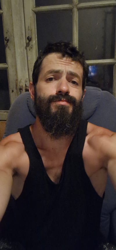 A man with a full beard and dark hair is taking a selfie. He is wearing a black tank top and is seated in a chair. Behind him, a wooden door with glass panes is visible, with a dark interior behind the glass. The man's left arm and shoulder are visible in the foreground. The scene appears to be indoors, likely in a residential setting. The image does not provide any information that would allow for the identification of the location as Wattenwil, Switzerland.