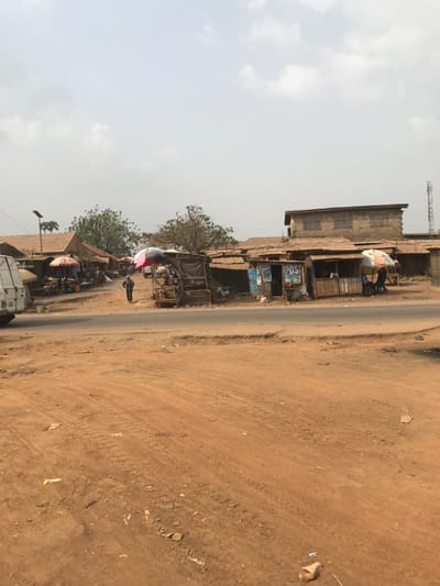 The image captures a bustling street scene in Ogbese, Nigeria, characterized by an unpaved reddish-brown earth foreground marked with tire tracks and minor debris. An asphalt road traverses the midground. Along both sides of this road, a series of single-story market stalls are visible, constructed with wooden frames and topped with corrugated metal roofs. Several stalls are shaded by open market umbrellas, featuring colors such as red and multi-striped patterns. On the right, at least two stalls bear prominent "POS" (Point of Sale) signage. Behind these stalls, a two-story concrete building with multiple windows is present, and further right, a metallic cellular communication tower is visible against the sky. On the left side of the road, a white commercial van is partially visible. Further back on the left, a solar-powered street light pole stands. Green trees are scattered throughout the background. Approximately five individuals are observed; one person walks centrally on the road section to the left, while others are situated near or within the market stalls, some appearing to be seated. The sky is overcast with a light grey hue, indicating daytime.