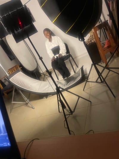 A young Black woman with dark hair pulled back is seated on a black chair in a photography studio. She is wearing a white collared shirt with a black sash or corset and black pants. She is smiling and looking towards the viewer. In the background, there are various photography lighting equipment including large softboxes, stands, and a large curved reflector. There is also a wooden stool and a section of a wooden shelf visible. The floor is tiled. The setting appears to be an indoor photography studio, likely located in South Ropp, Nigeria.