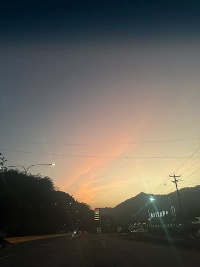 The image depicts a dusk or dawn scene near Cortada de Maturín, Venezuela. A highway runs alongside a hillside. Silhouetted trees and the elevated terrain are visible. Overhead power lines are suspended. The name "HAITANA" in illuminated letters is visible on the hillside. A PDVSA sign is also visible. Streetlights illuminate the roadway. The sky transitions from dark blue to lighter, yellowish-pink.
