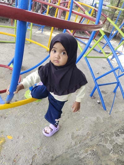 A young child wearing a black hijab and blue jeans is standing on a yellow bar of a playground structure in Kebunbaru, Indonesia. The child is looking directly at the camera with wide eyes. The playground structure is made of colorful metal bars in blue, red, green, and yellow. The ground is a concrete surface with scattered paint marks. The child's left hand is gripping a blue bar of the structure.