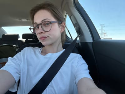 A young woman with glasses is in the passenger seat of a car, looking at the camera. She is wearing a white t-shirt and a seatbelt. The interior of the car is visible, including the back seats and the headrests. Outside the car, power lines and a tower are visible against a clear blue sky. The image was taken in Magnuszowice, Poland.
