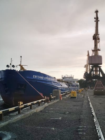 The image depicts a port scene in Shal'skii, Russia, under an overcast sky. In the foreground, a large blue cargo ship named "ЕВГЕНИЙ ОРЛОВСКИЙ" is docked alongside a pier. The ship is secured by a thick red rope. Adjacent to the ship are railway tracks laid on the ground, which is covered in gravel. Several yellow rectangular markers with numbers "5" and "6" are placed along the tracks. Two large cranes, one more prominent than the other, are visible in the background, suggesting loading or unloading operations. The environment appears industrial, with the water of the port visible on the left. There are no people visible in the frame. The lighting suggests it might be early morning or late afternoon.