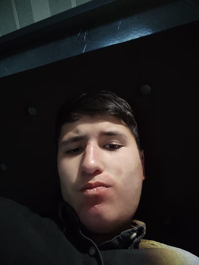 An indoor, low-light, close-up image depicts a young adult male, likely in Rishtan, Uzbekistan. The subject, with dark hair, dark eyes, and fair skin, is centrally positioned, looking directly at the camera. A reddish mark is observed on the bridge of his nose, and his lips are pursed. He wears a dark, collared shirt with light-colored buttons on the placket; a section of a lighter, patterned garment is visible on his right shoulder/chest area. The background is predominantly dark, featuring a dark, horizontal structure, possibly a furniture headboard, with two embedded circular, reflective elements. Above this structure, a vertically striped textile, possibly curtains, is partially visible in the upper left quadrant. Thin, light-colored linear abrasions are visible on the dark horizontal structure.