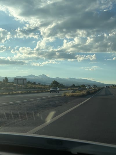 A wide-angle shot captures a highway with a mountainous landscape in the background. The sky is filled with dramatic clouds, some bright white and others dark gray, creating a dynamic contrast. In the middle ground, a highway stretches into the distance, with several cars visible on it. To the left of the highway, a large white sign stands beside a grassy embankment. The foreground shows the dashboard and windshield of a vehicle. The scene appears to be set in Proshyan, Armenia, likely during daylight hours, given the lighting conditions and presence of vehicles on the road.
