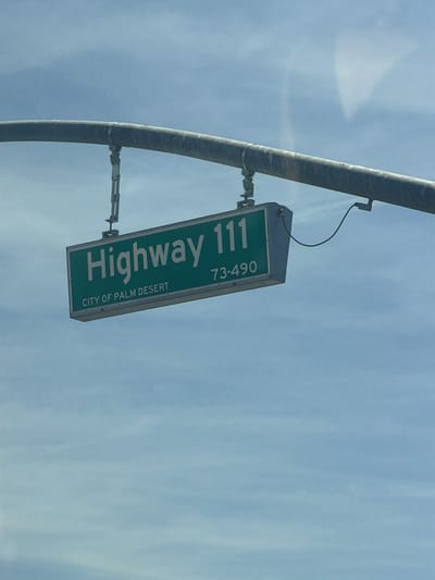 The image shows a green "Highway 111" sign with white lettering. Below that, it reads "CITY OF PALM DESERT" in smaller white lettering. The number range "73-490" is on the same line as "Highway 111", towards the right side of the sign. The sign is suspended from a curved, metallic pole by metal brackets. A thin black wire extends from the right side of the sign. The background is a bright blue sky with some light white clouds.