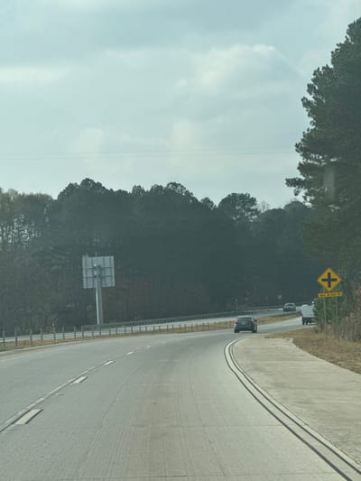 A highway curves slightly to the right, bordered by a metal guardrail. Trees line the left side, their leaves casting shadows. Two vehicles are visible on the road. A yellow road sign with a cross intersection symbol is also present. Further down the road, a large gray rectangular road sign is mounted. The sky is overcast. The image is slightly rotated.