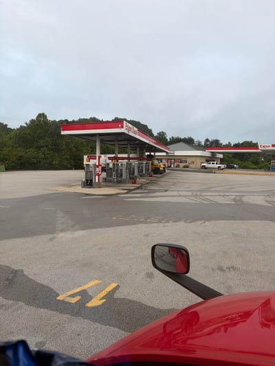 A red truck cab is in the foreground, with a side mirror reflecting part of the truck and the scene behind it. The truck is parked in an asphalt lot at an Exxon gas station in Wildwood, United States. Several gas pumps with red awnings and "Exxon Diesel" signage are visible. A yellow school bus, a white pickup truck, and two dark sedans are parked in the lot. In the background, a wooded area with green trees is present. The sky is overcast and cloudy. Yellow painted numbers "11" are visible on the asphalt in front of the truck.