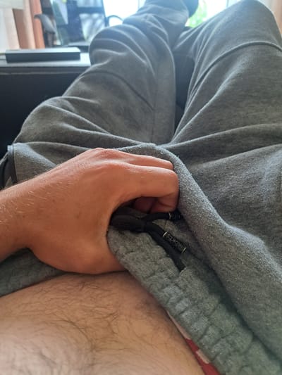 The image shows a close-up view of a person's hand gripping a grey fabric, possibly sweatpants or a hoodie. The fabric has drawstrings with black plastic adjusters. To the right of the hand, a portion of a leg covered in light hair is visible. In the background, a blurred window with greenery outside suggests an indoor setting, possibly a home, on a bright day. The scene is neutral in tone, with no discernible emotions or specific time of day, other than it appears to be daytime. No visible text is present in the image. The location context is Cēsis, Latvia.