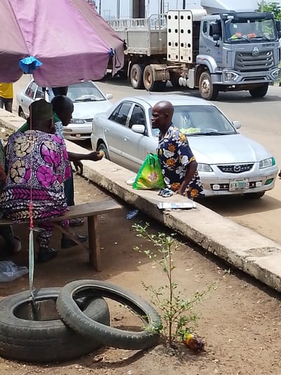 The image shows a street scene in Oja Oba, Oko Erin, Nigeria. In the foreground, a young plant grows next to a discarded tire. To the right, a concrete barrier runs along the side of the road. Behind the barrier, two men are interacting. One man, wearing a colorful patterned tunic and trousers, is seated on a wooden bench and holding a small yellow object in his right hand. He appears to be in conversation with the second man, who is standing and wearing a short-sleeved shirt with a floral pattern. A green plastic bag is placed on the ground near the standing man. In the background, a silver sedan car is parked on the road, with its license plate visible as "AFN 700AE". Further in the background, a large silver truck is also visible on the road. The scene is partially shaded by a large purple umbrella. The ground is a mix of dirt and scattered debris.