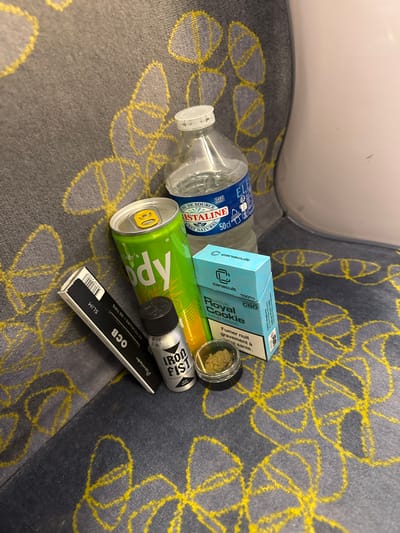 A collection of personal items is scattered on a patterned grey and yellow fabric surface, likely a seat in a public transport vehicle or waiting area. The background features a light-colored, curved interior surface.

The objects include an almost empty transparent plastic water bottle, labeled 'ISTALINE', 'EAU DE SOURCE', 'ETAT NATUREL', and '50cl', indicating natural spring water without nitrates. Next to it, a light green aluminum can with a yellow smiley face tab is partially visible, bearing text like 'Cody's' and possibly 'Xyro'.

A light blue box, branded 'canacult', 'SATIVA Royal Cookie', is present, stating '100% CBD' and 'SANS TABAC' (without tobacco), along with the warning 'Fumer nuit gravement à santé' (Smoking seriously harms health). A small, clear glass jar containing what appears to be ground plant material, consistent with CBD flower, sits beside the box.

Further to the right, a small silver and black cylindrical object with text 'IRON FIST' and dates '1698', '07/027' is visible. Below these items lies a black packet of rolling papers, 'OCB Premium SLIM', specifying '32 FEUILLES + 32 TIPS'.

The arrangement of these items suggests recent use or preparation for consumption, particularly of the CBD product, possibly by rolling it. The French text on several items is consistent with the provided location context of Orsay, France, although no specific landmarks are visible.