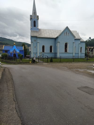 This image depicts a street scene in Bohdan, Ukraine, with a light blue church prominently featured on the right. The church has a tall, pointed steeple and multiple arched windows. To the left of the main church, a smaller, brightly colored blue building with a golden dome is visible, possibly another religious structure or a chapel. A black metal fence encloses the area around the blue church, and a person on a bicycle is positioned near the entrance gate. The road in the foreground is paved and appears wet, suggesting recent rain. Hills covered in dense green trees form the background, under a cloudy sky. Power lines are strung across the upper portion of the image.