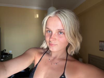 This media file is a close-up, upside-down, portrait-style photograph of a young woman. She has fair skin with freckles, light blonde hair that is styled in soft waves, and light-colored eyes. She is wearing a black bikini top adorned with silver grommets and a delicate silver necklace with small beads. The setting appears to be indoors, with a warm, yellow-toned wall and white baseboards visible in the background. Sunlight is casting a rectangular reflection on the wall to the right of the subject's head. The lighting suggests it might be daytime, possibly late afternoon, given the soft quality of the light. The woman's expression is neutral, and the image is taken from a selfie perspective. No text is visible in the image. The location context provided is Arkadii Municipal Unit, Greece.