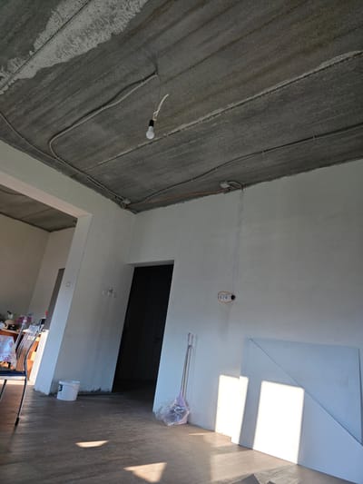 The image depicts the interior of a room under renovation in Bolshiye Kharlushi, Russia. The room features a raw concrete ceiling with visible electrical conduit and a single bare lightbulb hanging by a wire. The walls are painted white, with some showing signs of unfinished plastering or staining. There is a dark doorway on the right side of the image, and another opening on the left leading to an adjacent area. Several objects are scattered around the room, including a white bucket near the left doorway, a broom with pink bristles partially wrapped in plastic, and two large white sheets of material leaning against the right wall. The floor is a dark wood laminate. Sunlight streams in from an unseen window, casting distinct shadows across the floor and onto the white sheets. There are no people visible in the scene.