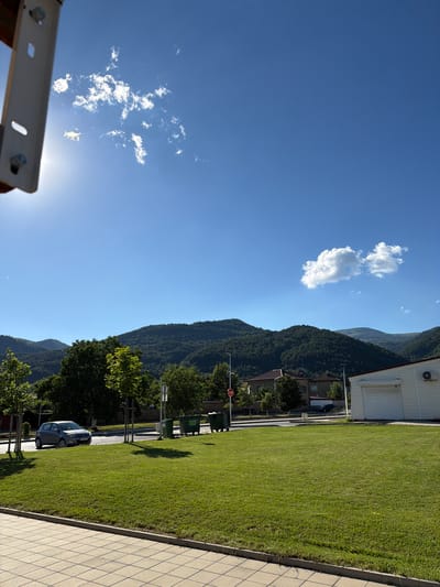 The image shows a sunny day scene in Chelopech, Bulgaria. In the foreground, there is a manicured green lawn bordered by a tiled pathway. Beyond the lawn, a road runs horizontally, with a gray sedan driving on it. Across the road, there are several green trash bins and some trees. In the middle ground, residential buildings with red tile roofs are visible, nestled among trees. Further in the background, a lush, forested mountain range stretches across the horizon under a clear blue sky dotted with a few white clouds. The sun is bright, casting shadows and creating lens flare in the upper left corner.