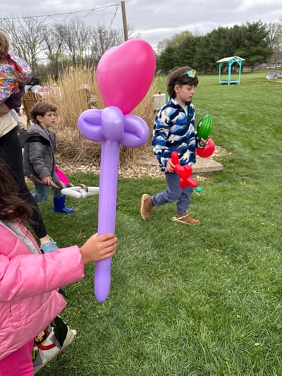 The image shows a group of children in an outdoor setting, likely in Pickerington, United States, as that information was given. One child is running across a grassy lawn, wearing a blue camouflage patterned jacket, blue pants, and brown boots. The child also has a red helmet, a green ball or small helmet, and a red balloon. Another group of children are near the lower edge of the image, with a girl in a pink jacket holding a large, purple balloon creation. There are bare trees and shrubs in the background. A small blue and white gazebo is visible on the lawn behind the running child.