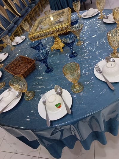 A decorated round table is covered with a blue vinyl tablecloth. Place settings include white plates with small bowls turned upside down, forks, and spoons. Each setting also has a decorative glass, some in blue and others in gold. A brown wicker basket sits near one setting. An ornate gold-colored centerpiece is placed near the center of the table. Blue-upholstered chairs line the background.