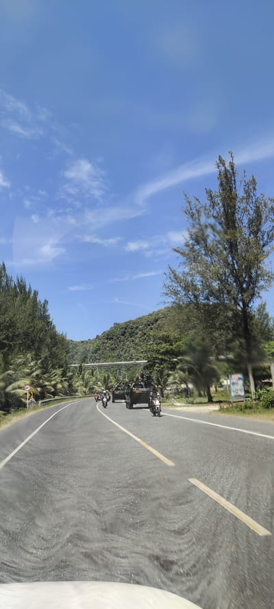 In Lamkruet, Indonesia, a convoy of military vehicles, including an armored personnel carrier and several motorcycles, travels along a paved road. The road is bordered by lush green vegetation, including palm trees and a dense forest covering a hilly landscape. The sky above is a clear blue, with scattered white clouds. The convoy appears to be in motion, suggesting a patrol or transit operation. The scene is set in a tropical environment, with the presence of palm trees indicating a coastal or warmer climate.