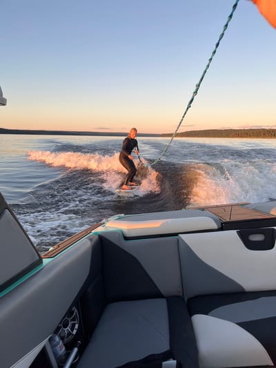 A blonde woman in a black wetsuit is wakeboarding behind a boat on a lake at sunset. She is holding onto a tow rope and riding on a wave created by the boat. The boat is in the foreground, and the wakeboarder is in the middle ground. The background shows a treeline and a clear sky with sunset colors. The image appears to have been taken from another boat, looking towards the wakeboarder. The location is specified as Gromovo, Russia.