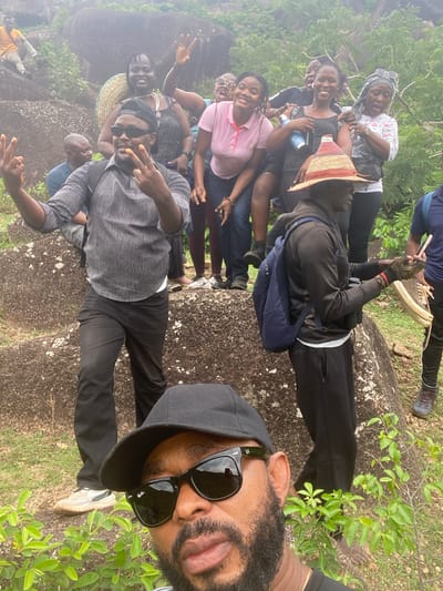 A group of people are posing for a photograph on what appears to be a rock formation, possibly in a rural or natural area near Ganawuri, Nigeria. The group includes men and women of various ages. They are dressed in casual clothing. Some are smiling and making peace signs. A man in the foreground wears sunglasses and a black baseball cap. The environment includes rocks and green vegetation. The background is slightly blurry, possibly due to the depth of field.