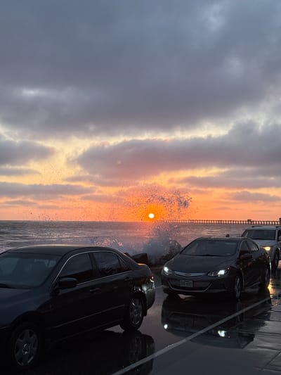 The image captures a sunset at the beach with rough ocean waves crashing against the shore. A pier is visible in the distance. In the foreground, several cars are parked on a wet surface, likely a road or parking lot adjacent to the beach. The sky is filled with dramatic clouds illuminated by the setting sun, casting orange and pink hues. The overall scene depicts a stormy sunset by the coast.