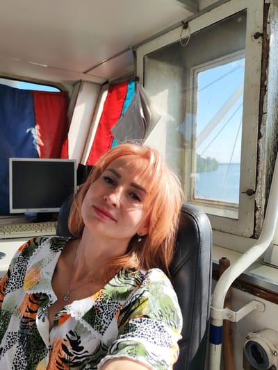 A woman with reddish-blonde hair and makeup is sitting in a chair inside what appears to be a boat's cabin or control room. She is looking towards the camera with a slight smile. She is wearing a colorful, patterned shirt with a V-neck and a silver necklace with a pendant. Her hair is styled with bangs. She has earrings on.
Behind her, to the left, is a computer monitor and keyboard. The monitor screen is black. There are flags hanging on the wall behind the computer. One flag appears to be blue and red, and another is red.
To the right of the woman is a window that looks out onto a body of water with trees visible on the distant shore. The sky is blue. The sunlight is coming in from the right, casting shadows. The interior of the cabin is utilitarian, with white walls and metal elements. There is a white pipe with a blue band running along the right side of the image.
The scene suggests a daytime setting, likely sunny given the lighting. The presence of the water, flags, and the cabin's interior points to a location on a body of water, possibly a river or lake in Nova, Russia, given the context. The woman's relaxed pose and smile might indicate she is comfortable and perhaps taking a break.
