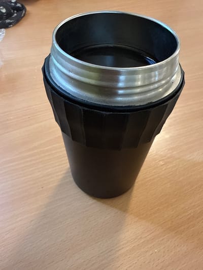 A black thermos with a silver rim is sitting on a light-colored wooden surface. The thermos is open at the top and appears to contain a dark liquid. The background shows a blurred object with a black and white pattern. The scene is well-lit, casting a shadow of the thermos onto the wooden surface. There are no people present in the image. The location of Södra Svartbyn, Sweden is not depicted or suggested by the image content.