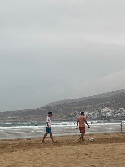 The image shows two people playing with a soccer ball on a beach in Taghazout, Morocco. The beach is sandy and slopes down to the ocean, which is moderately wavy. A town is built into a hillside that rises to the right of the beach. The sky is overcast. One person is wearing a salmon-colored shirt and salmon-colored shorts, and is facing the soccer ball. The other person is wearing a white shirt and blue shorts, and is looking toward the first person and the soccer ball. They are both barefoot.