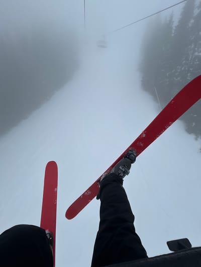 The photograph captures a skier riding a ski lift in foggy conditions. The skier is wearing black ski pants and boots, attached to red skis covered with light snow. The view is from the skier's perspective, looking down at the skis and the foggy landscape below. A ski lift tower is visible through the fog in the distance. The location is Stoikite, Bulgaria.