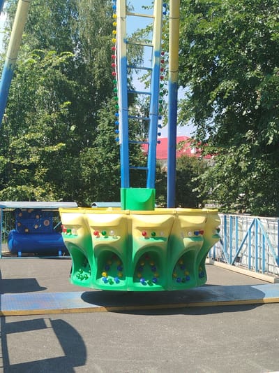 The image shows a children's amusement ride in an outdoor setting. The ride features a central structure with yellow and green elements, and a circular platform at its base containing colorful balls. In the background, there are trees, a blue tank, and a building with a red roof. The scene appears to be located in the city of Yalutorovsk, Russia, as indicated by the context provided. The ground is asphalt, and there are shadows suggesting it is daytime. There are no people visible in the image.